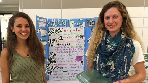 two female students standing in front of a poster, holding a green to-go box